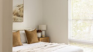 A bedroom with a bed, white bedding, a nightstand with a lamp and a vase, and a painting on the wall.