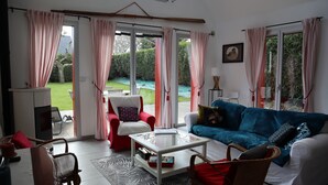 Una sala de estar con un sofá azul, un sillón rojo y una mesa de centro blanca. Hay ventanas amplias con cortinas, un televisor y un cuadro en la pared.