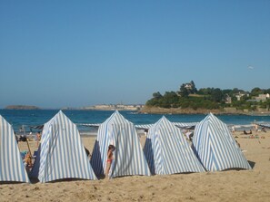 Gestreifte Strandzelte an einem Sandstrand mit freiem Blick auf das Meer und ein Gebäude in der Ferne.