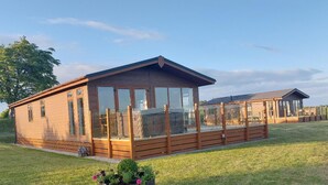 A wooden cabin with large windows and a deck, surrounded by grass and trees. - 2 Bedroom Sleeps 4 Lodge-smart Tv-hot Tub-dogs Ok (Lochwinnoch)