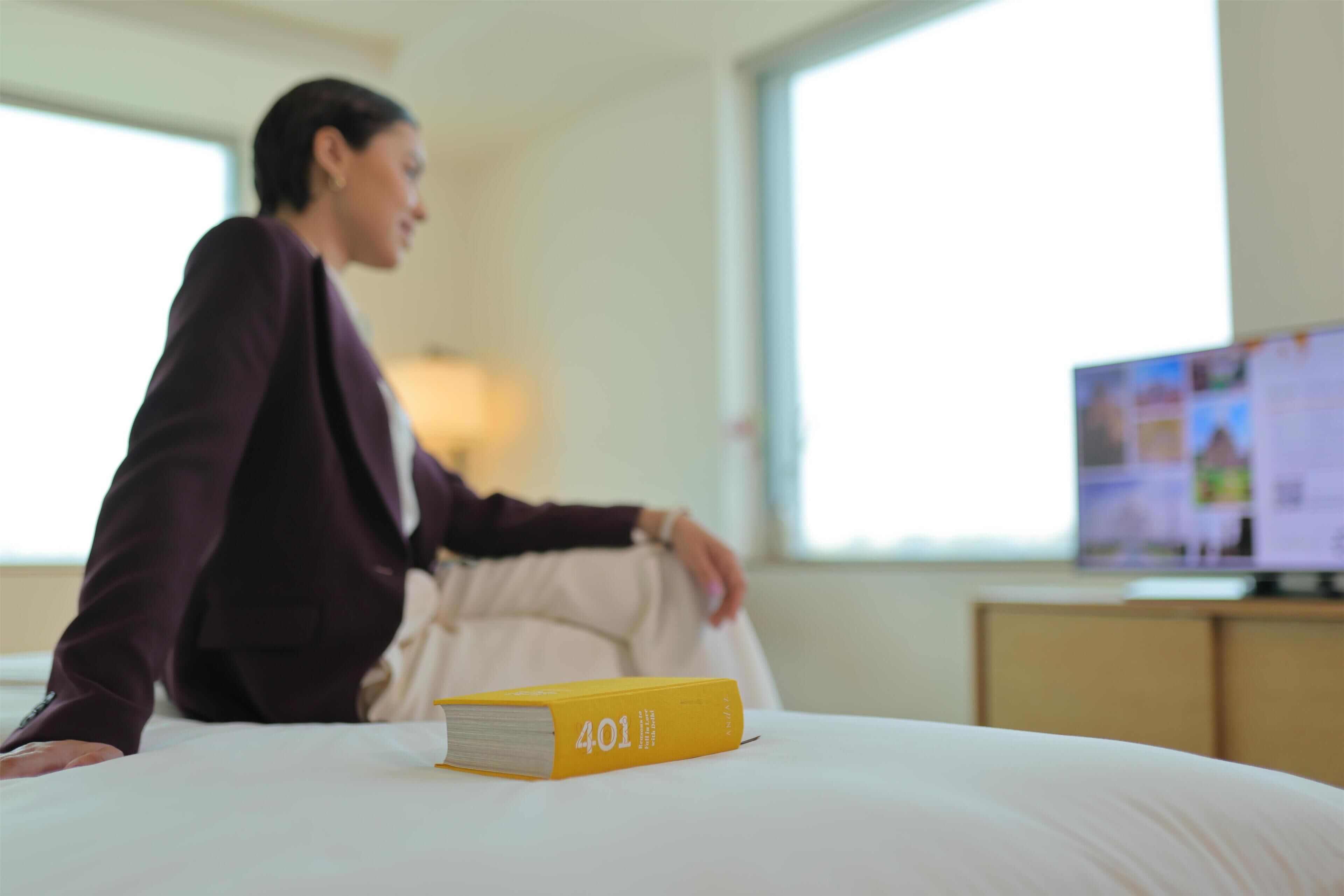 Eine Person sitzt auf einem Bett, ein Buch mit dem Titel '401' liegt daneben, im Hintergrund steht ein Fernseher.