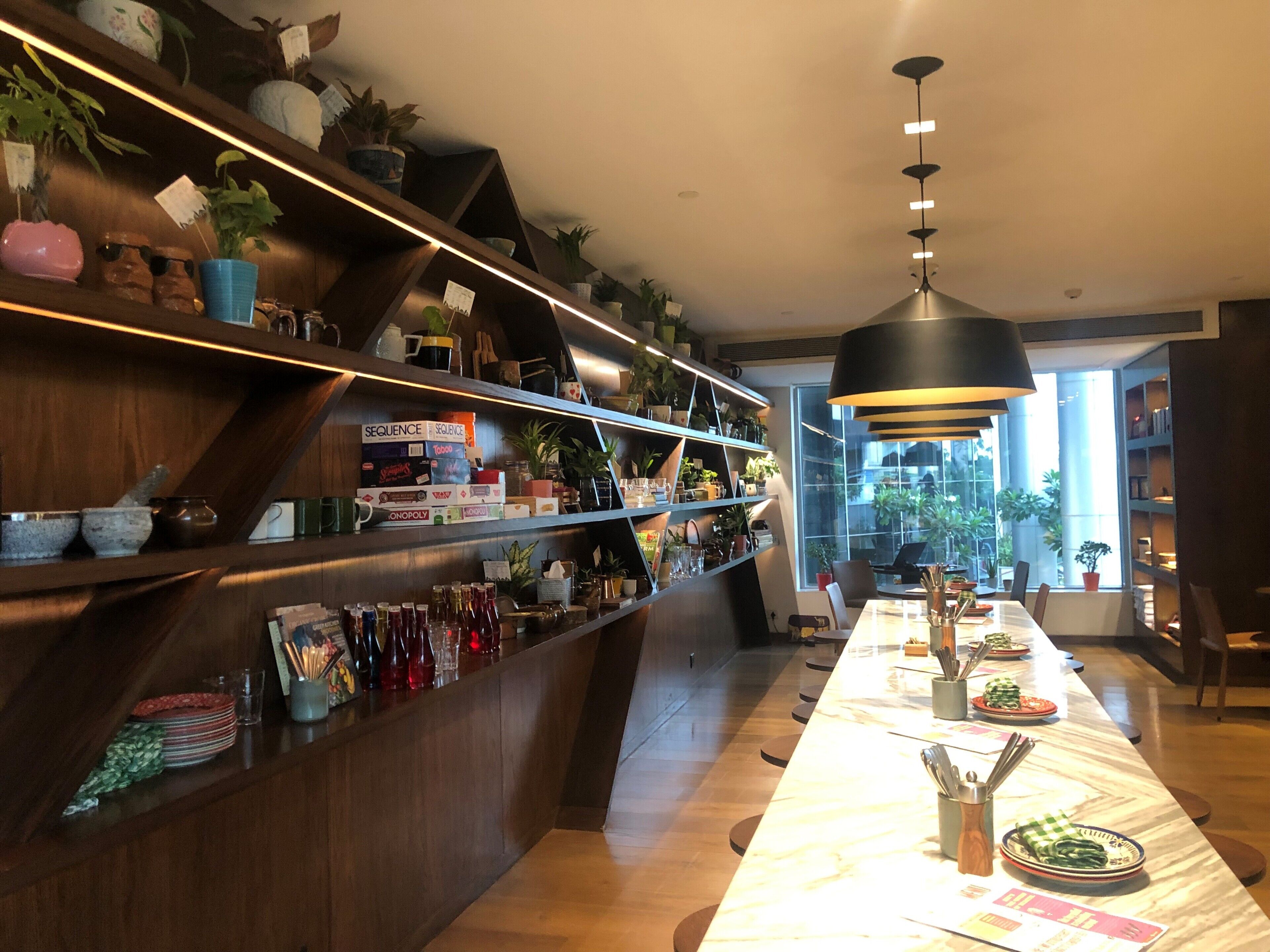 A modern restaurant with a long dining table, shelves with plants and kitchenware, and a large window providing a view of the outside.