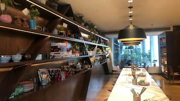 A modern restaurant with a long dining table, shelves with plants and kitchenware, and a large window providing a view of the outside.