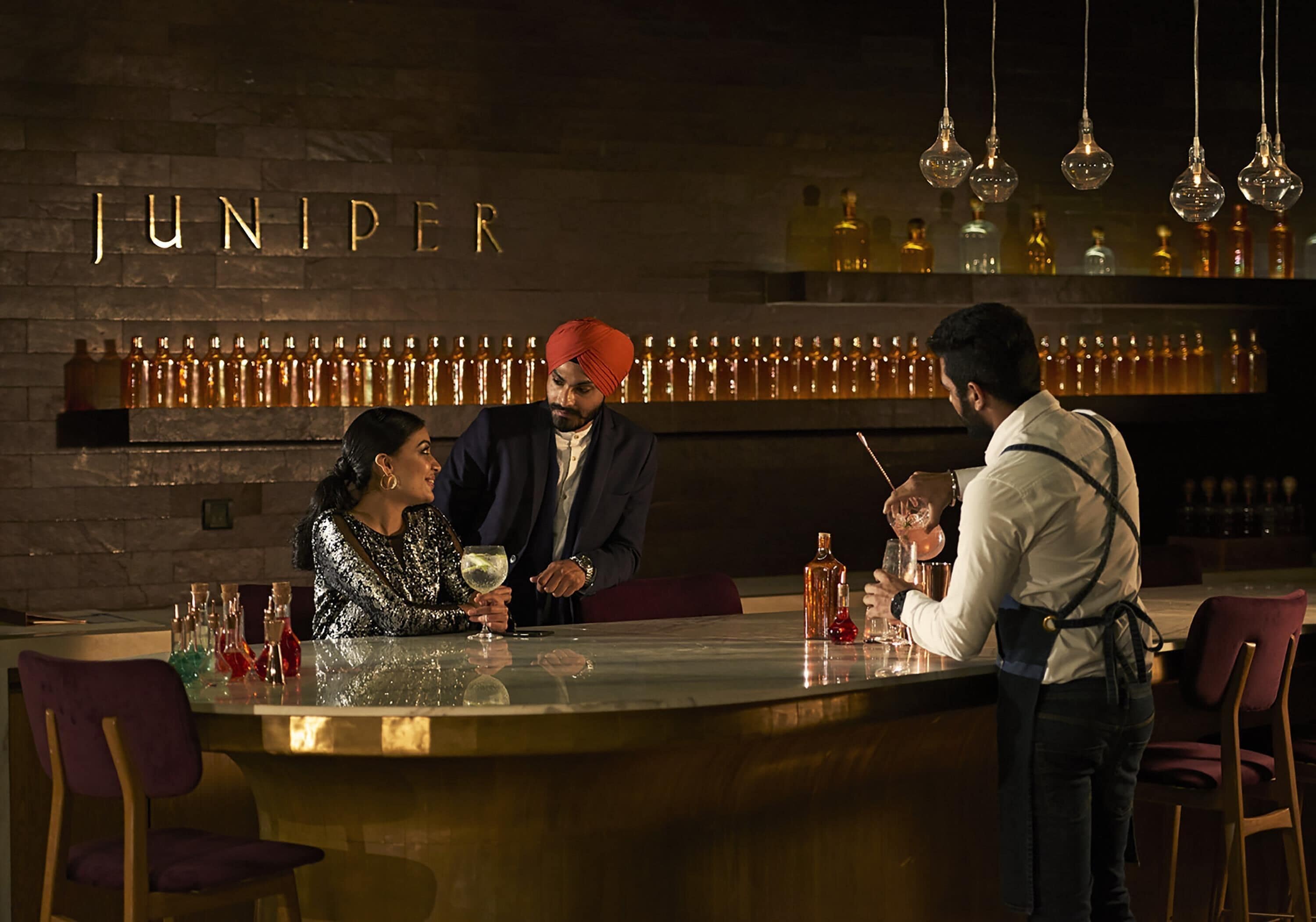 A bar area with a sign reading 'JUNIPER,' bottles of liquor, and a person serving drinks.