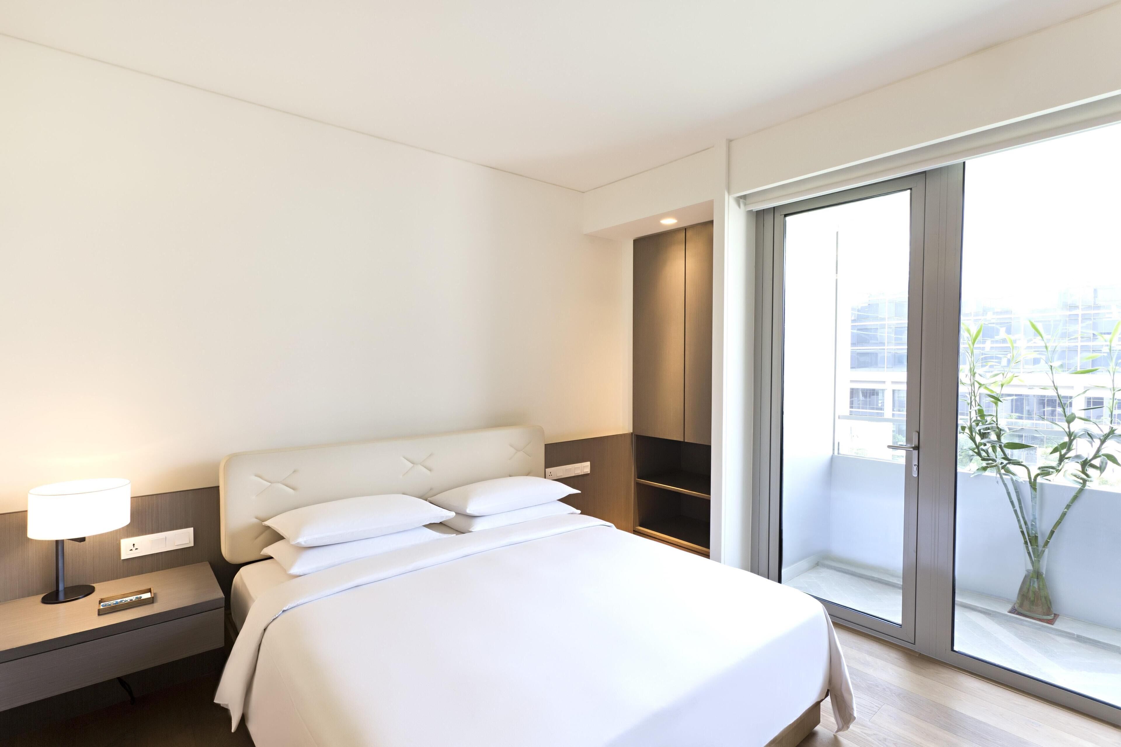 Habitación de hotel con cama, mesita de noche, lámpara y un ventanal con vistas a la ciudad.