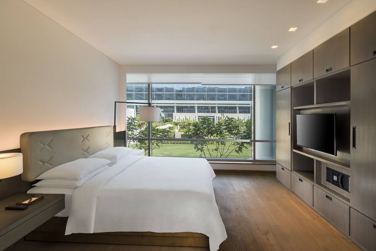 A modern hotel room with a large bed, a TV, and a view of a building and greenery outside.