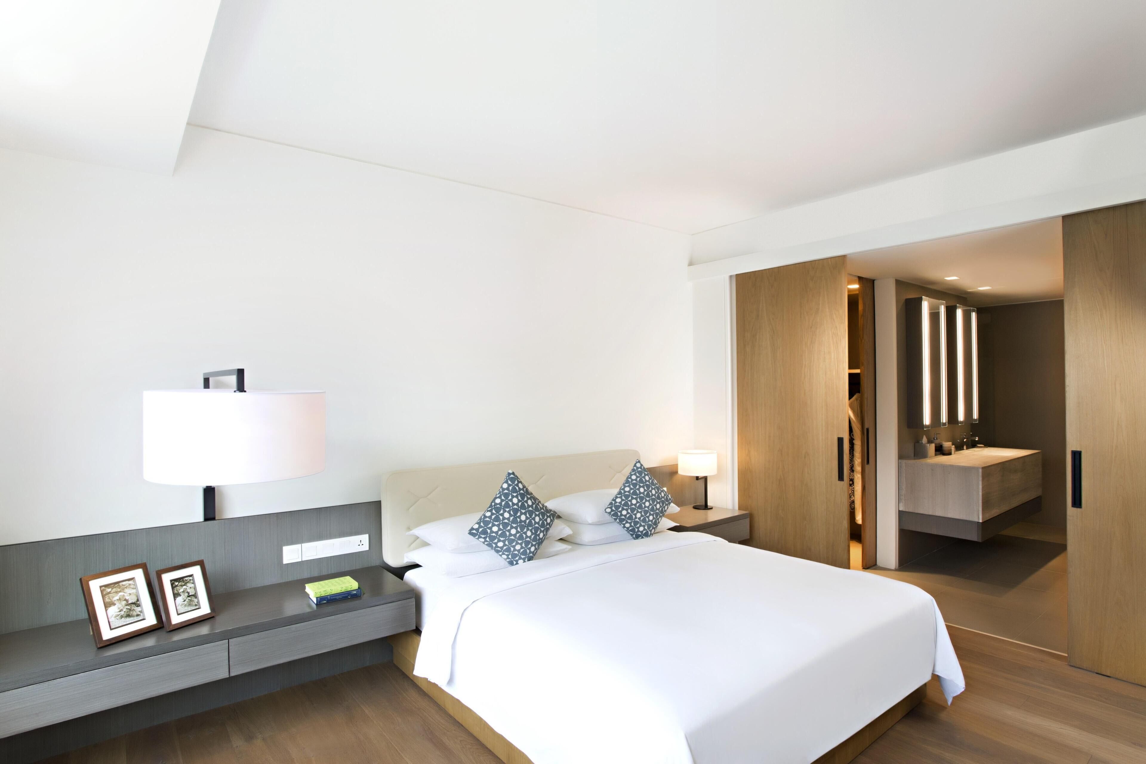 A modern hotel room with a bed, bedside tables, a nightstand, a mirror, and a bathroom visible through an open door.
