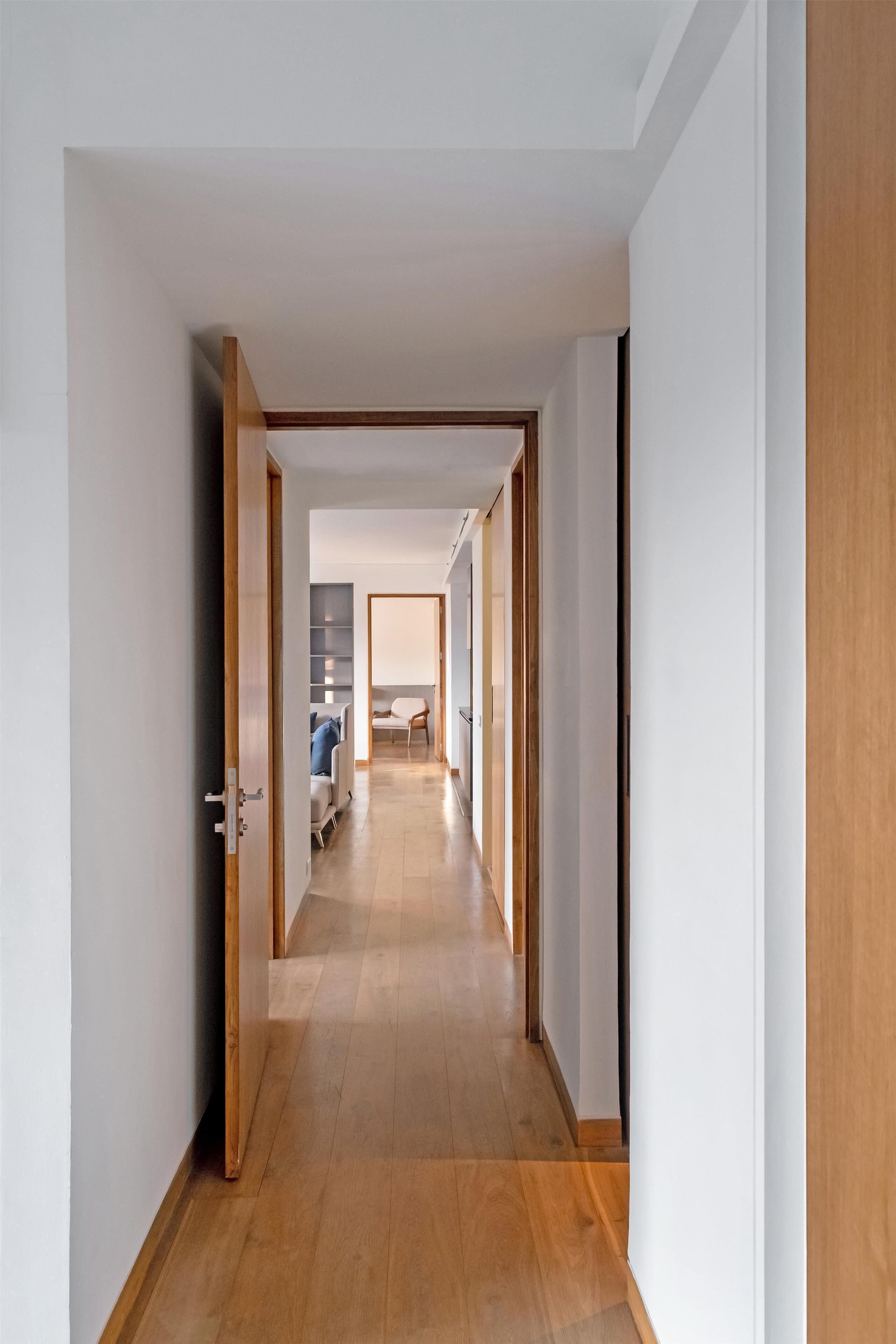 A hallway with wooden flooring, white walls, and a door leading to a room with a bed and a chair.