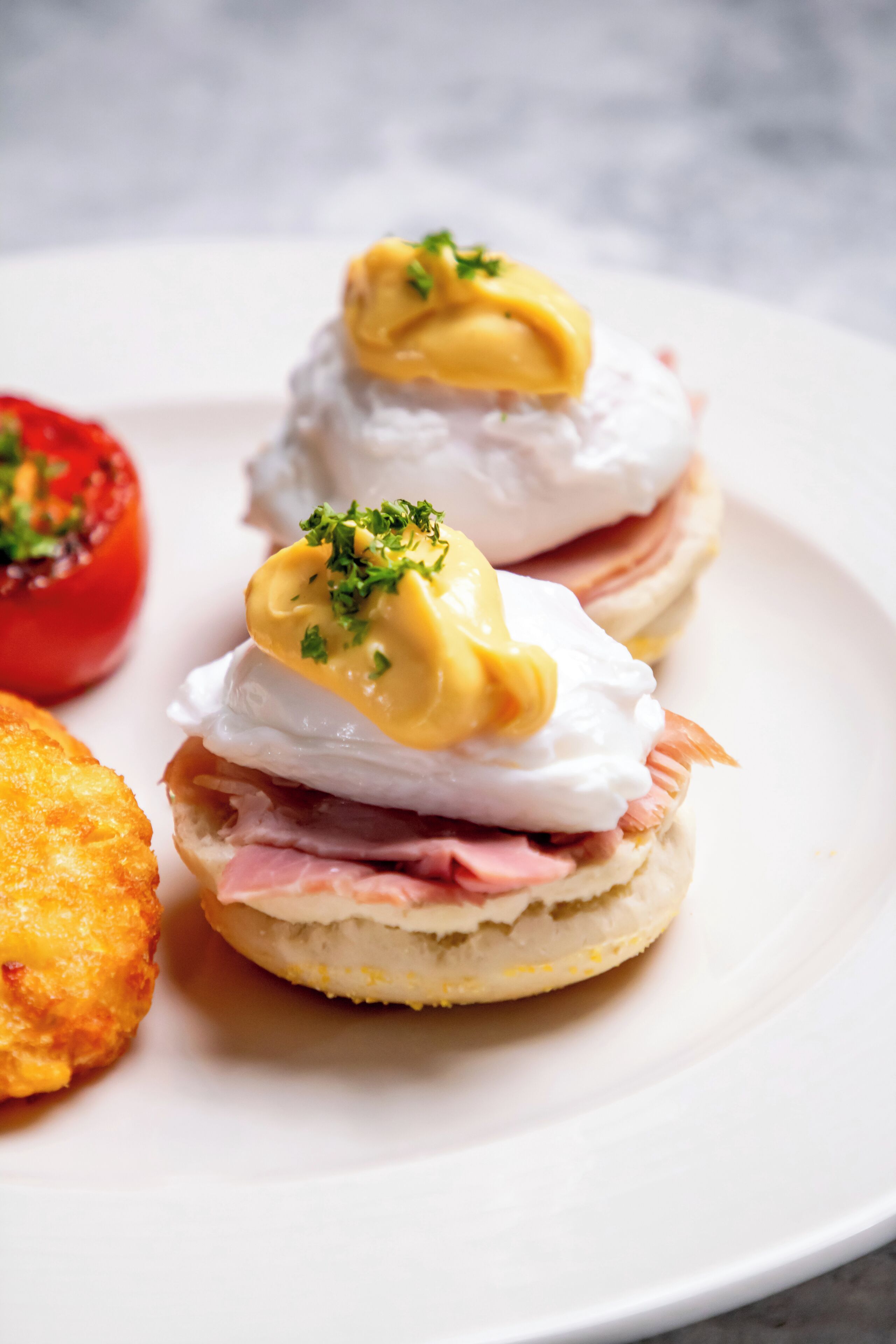 Une assiette avec deux pains anglais garnis de jambon, d’œufs pochés et de sauce hollandaise, décorée de persil, accompagnée d’une tomate rôtie.