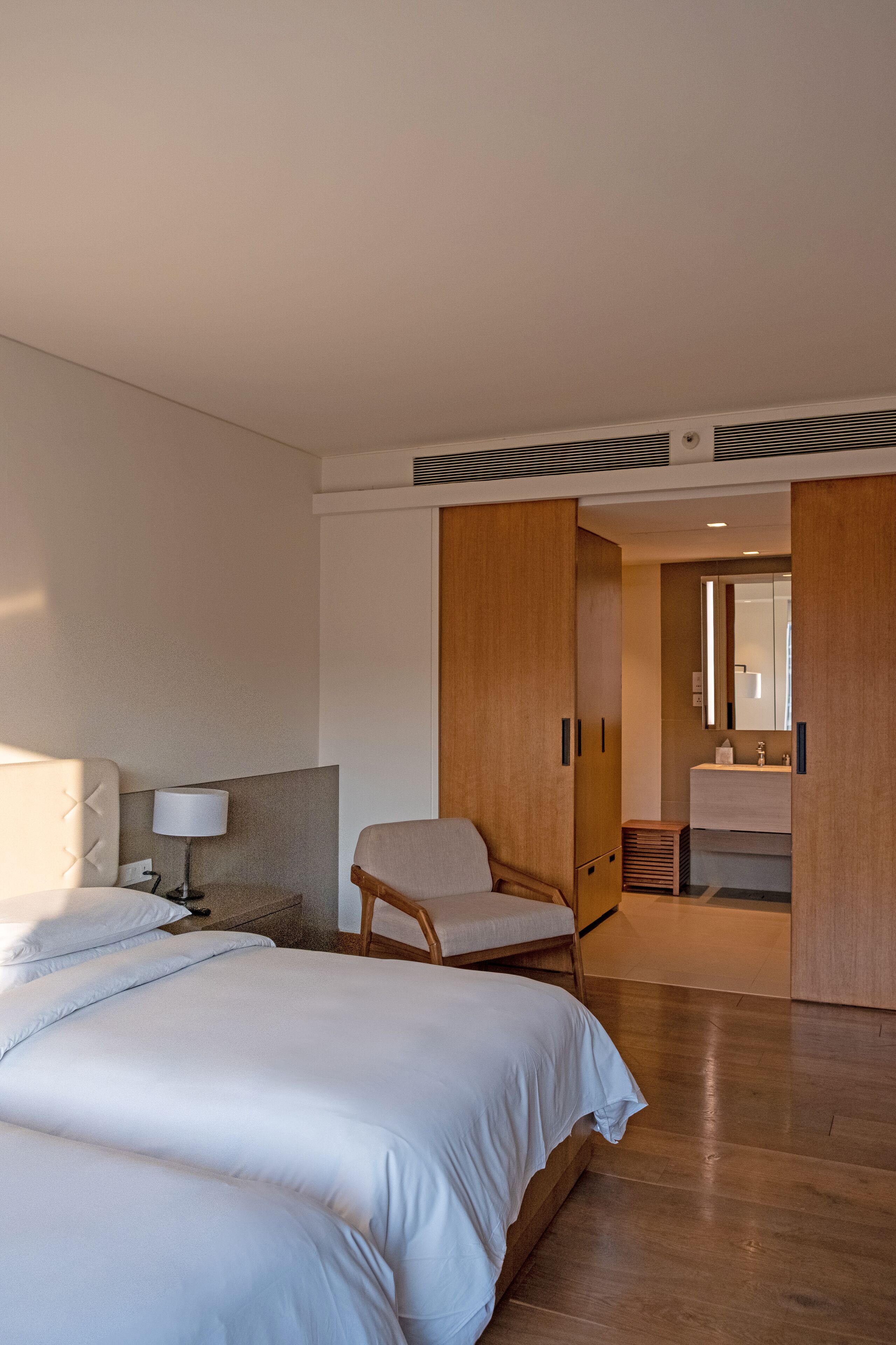 A hotel room with a large bed, a chair, a desk with a lamp, and a bathroom visible through an open door.