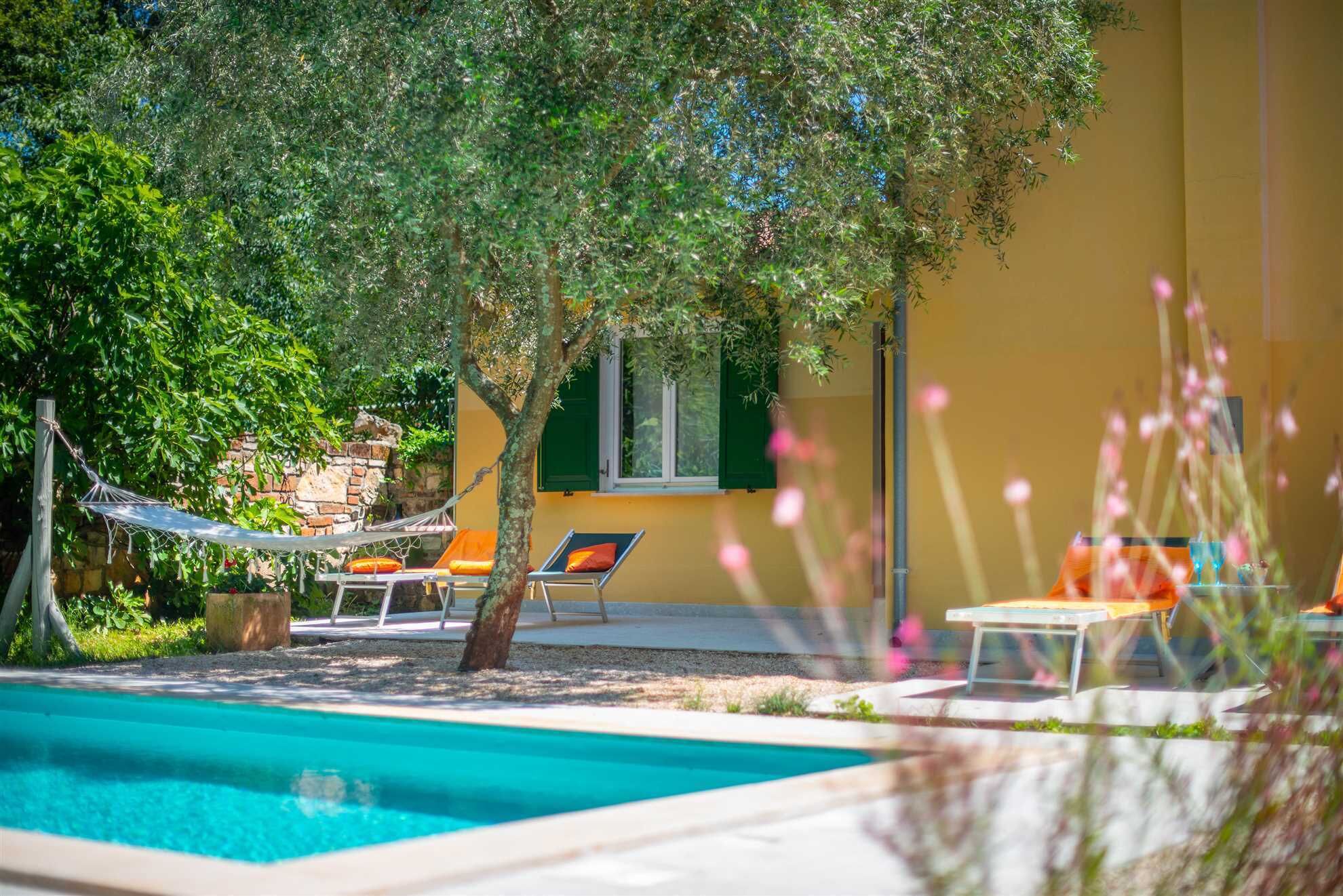 Un área de piscina con sillones de descanso y una hamaca, rodeada de follaje y un edificio amarillo.
