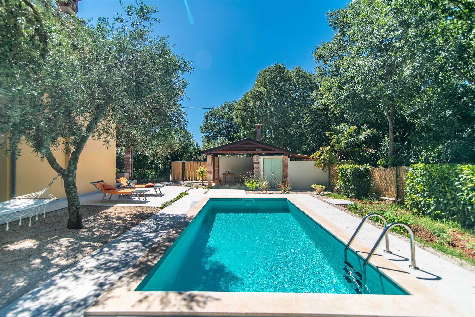 Piscina rectangular con cubierta de madera, rodeada de árboles y un pequeño edificio de techo rojo.