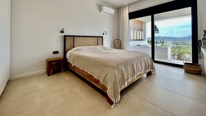 Une chambre dotée d’un grand lit, d’une table de chevet, d’une chaise et offrant une vue sur l’extérieur grâce à une porte-fenêtre coulissante.
