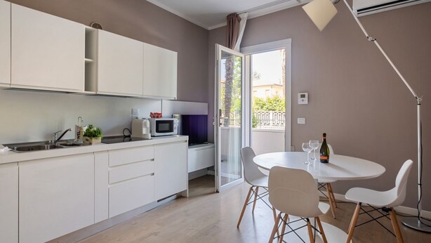 Een moderne keuken met witte kasten, een magnetron en een gootsteen. Er is een eethoek met een witte tafel en stoelen, en een balkon met uitzicht op gebouwen.