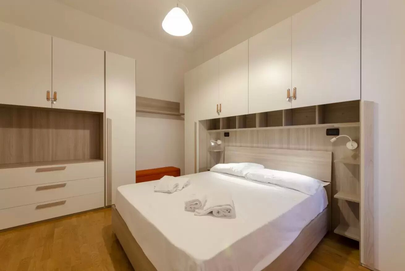 A modern hotel room with a large bed, white wardrobe, and a bedside table.