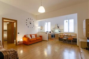 Amplia sala de estar con sofá, una mesa de comedor con sillas, zona de cocina y un reloj de pared grande.