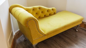 Um sofá amarelo com detalhes em tecido e uma corrente decorativa na lateral, posicionado em frente a uma parede branca em um quarto com piso de madeira.