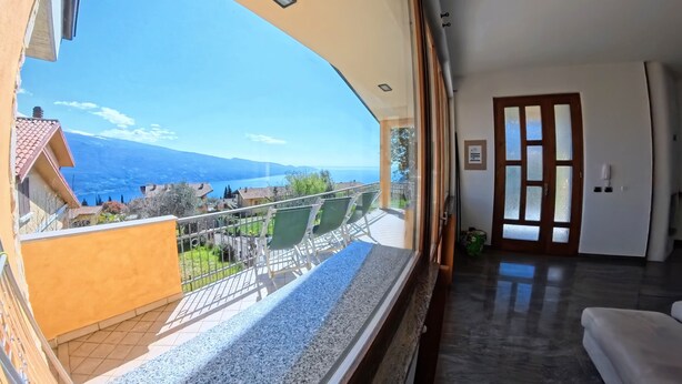 A room with a large window overlooking a balcony with a view of mountains and a lake. - Villa Arianna – Panorama Suite, Lake Garda view apartment with pool (Tignale)
