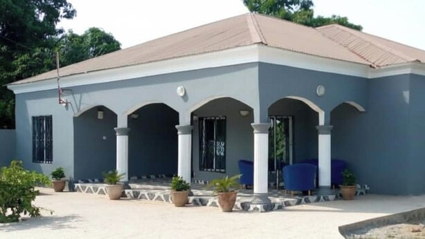 A two-story building with a light blue facade, white columns, and a red roof. - 4 Bed House Sleeps 8 - Garden and Pets Welcome (Brikama)