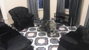 A living room with a patterned floor, a black armchair, a glass coffee table, and a TV stand. - 4 Bed House Sleeps 8 - Garden and Pets Welcome (Brikama)
