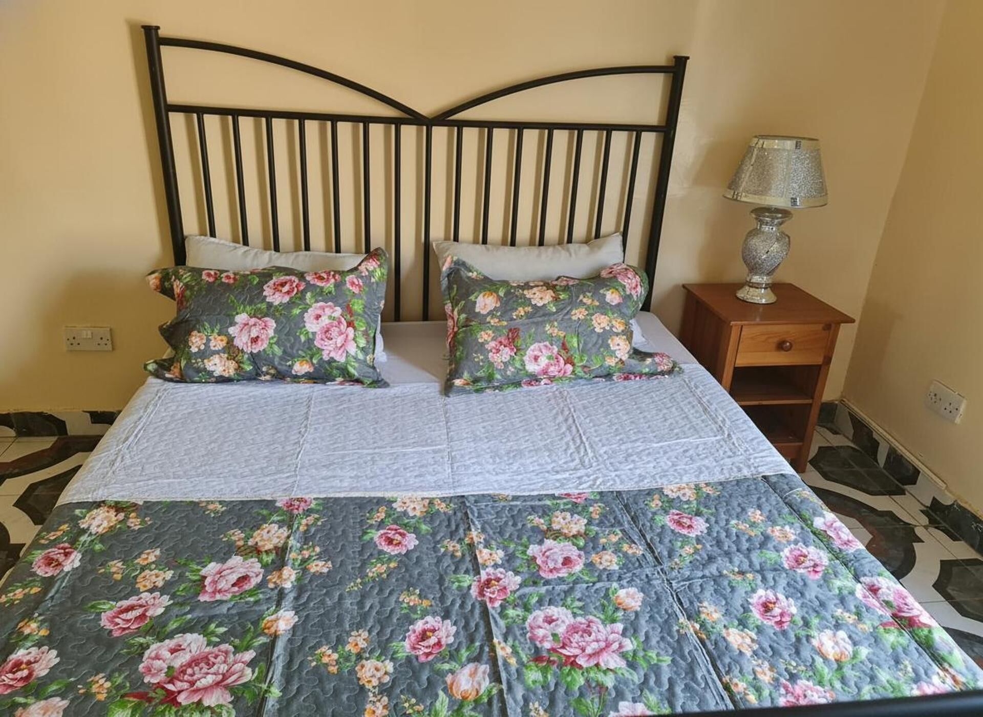 Una cama con un edredón floral y dos almohadas a juego, una mesita de noche con lámpara, y un suelo estampado.