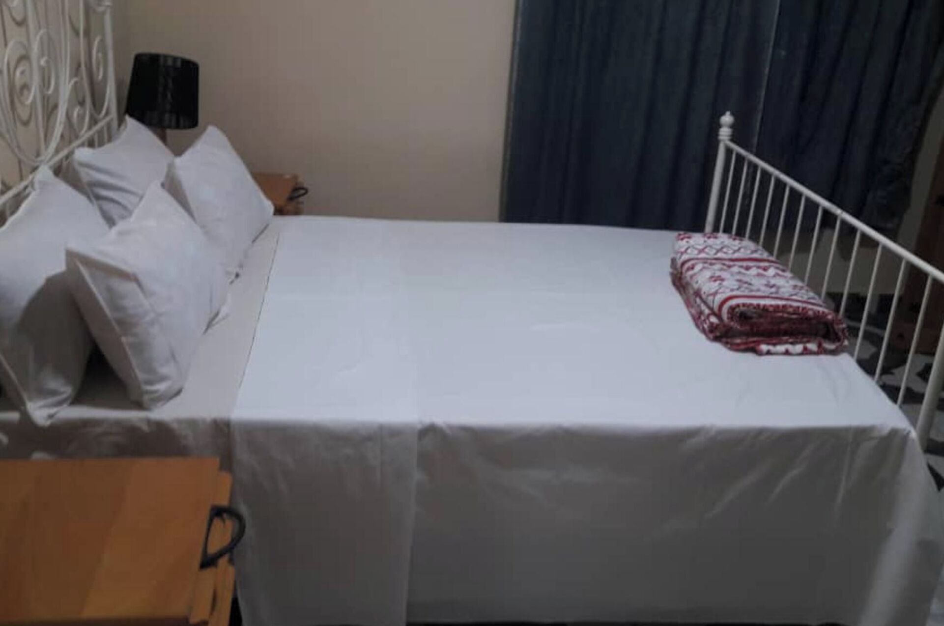 A single bed with a white mattress and a patterned blanket, a wooden nightstand, a lamp, and a curtain.
