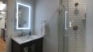 Une salle de bain avec un lavabo blanc, un miroir, une cabine de douche en verre et un meuble en bois foncé.