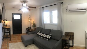 Un salon avec un canapé gris, une petite table, un ventilateur de plafond, un téléviseur fixé au mur et un appareil de climatisation.