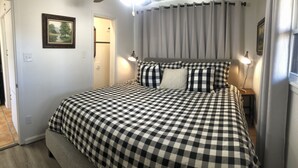 Une chambre avec une couvre-lit à carreaux, une tête de lit agrémentée de rideaux, une lampe de chevet et un tableau encadré accroché au mur.