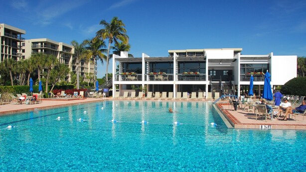 A swimming pool with clear blue water, surrounded by lounge chairs and umbrellas, and a modern building with balconies in the background. - Full Beachfront 2 BR Condo (Longboat Key)