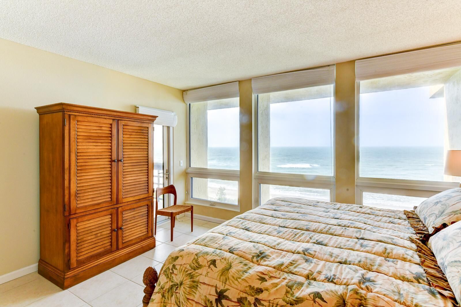 A bedroom with a large bed, a wooden wardrobe, a chair, and a view of the ocean through large windows.
