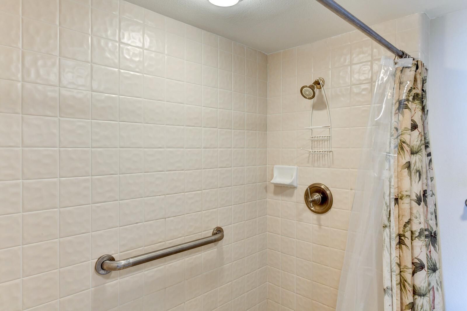 A bathroom with beige tiled walls, a shower with a curtain, a handrail, and a small shelf with a soap dish.