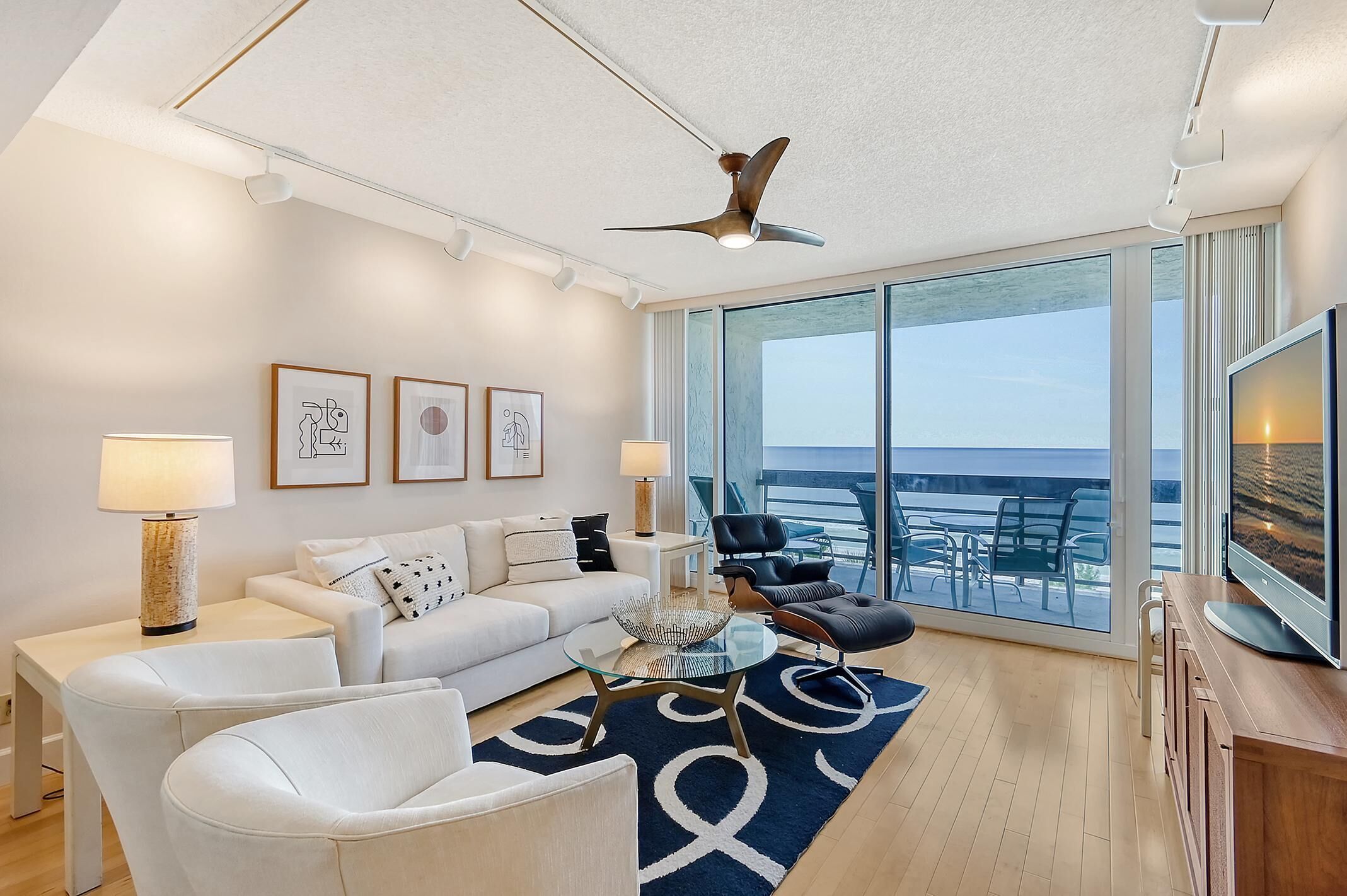 A modern living room with a sofa, armchairs, a glass coffee table, and a ceiling fan. There are framed pictures on the wall, a TV, and a large window with a view of the ocean.
