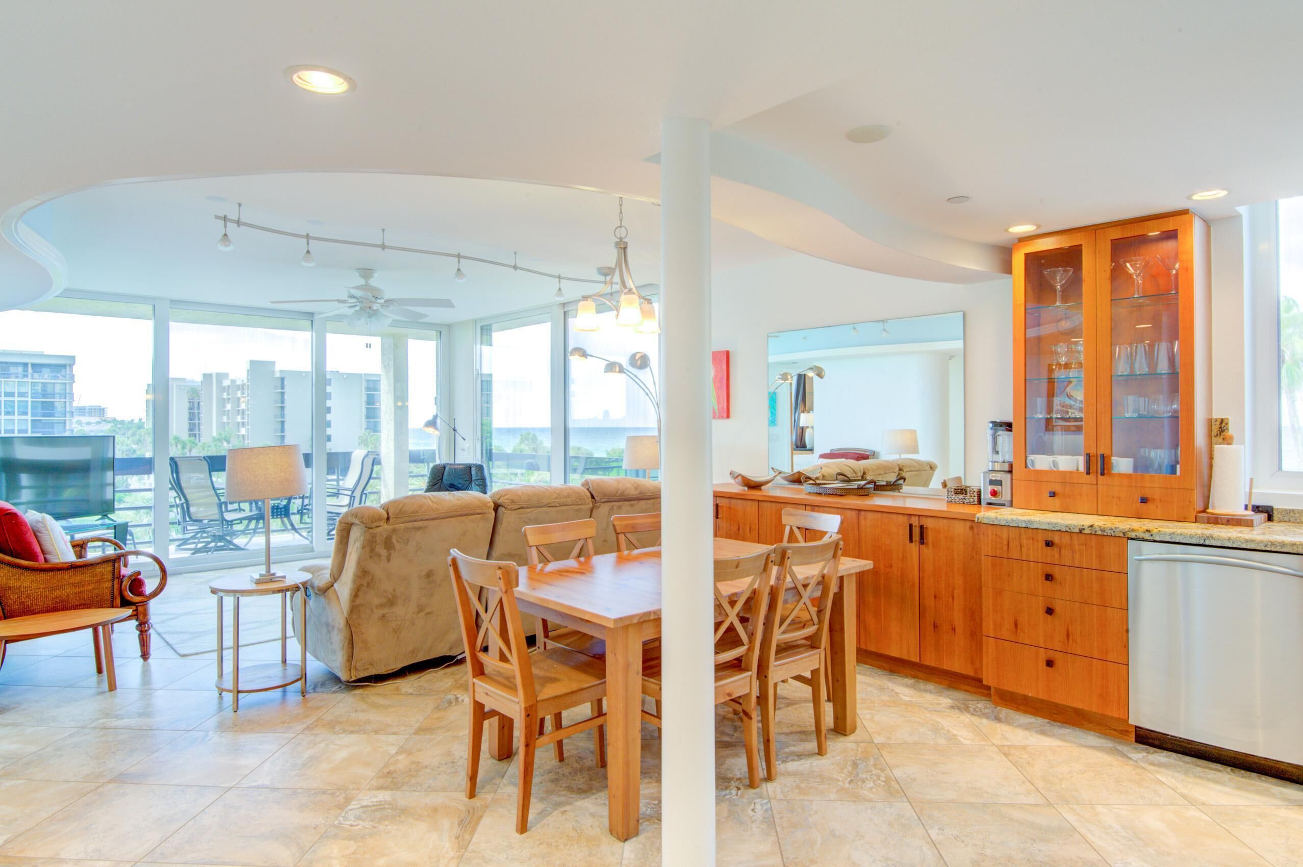 A spacious living area with a sofa, dining table, and chairs, a kitchen with wooden cabinets, and large windows offering a city view.