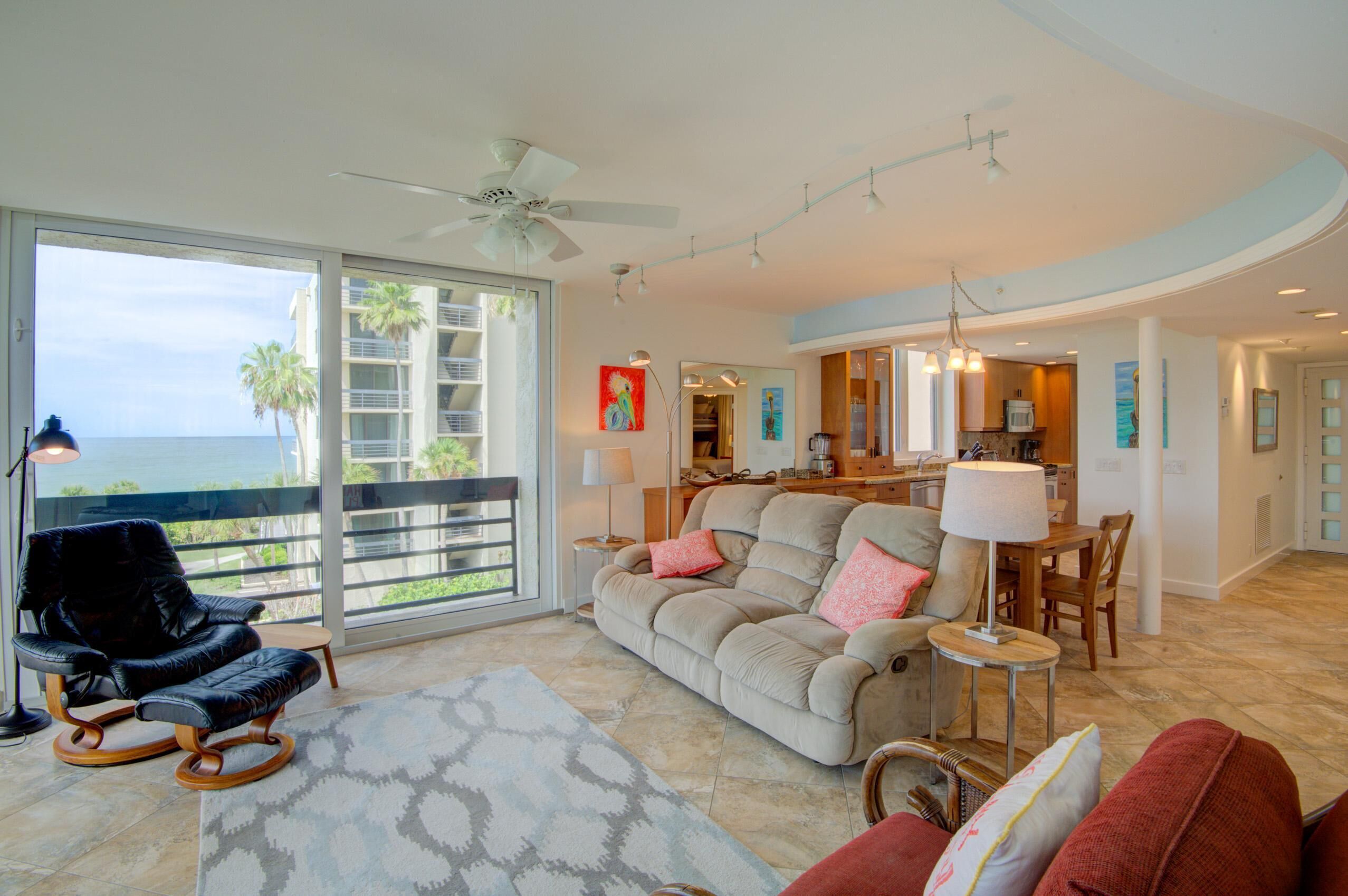 A spacious living area with a large window overlooking the ocean, a ceiling fan, and a dining area with a table and chairs.