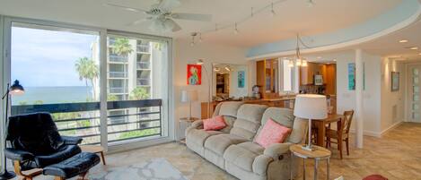 A spacious living area with a large window overlooking the ocean, a ceiling fan, and a dining area with a table and chairs.