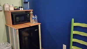 Eine kleine Küchenzeile mit Mikrowelle, Kaffeemaschine und Wasserflaschen. Ein kleiner Mülleimer ist links zu sehen.