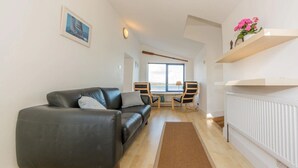 A modern living room with a black leather sofa, a wooden coffee table, and two chairs. A large window offers a view of the outdoors. - Mulroe Cove - The Boathouse, Fisherman's Lane (Mulroe)