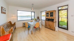 A dining area with a table and chairs, a wooden bench, a cabinet, and a view of the sea through large windows. - Mulroe Cove - The Boathouse, Fisherman's Lane (Mulroe)