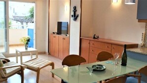 Un salon moderne avec une table à manger au plateau en verre, des chaises en bois, un canapé et un petit coin cuisine équipé de placards et d’un téléviseur.