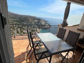 A balcony with a glass table and chairs, overlooking a coastal landscape.