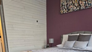 A bedroom with a bed, a bedside table, a lamp, and a painting on the wall. - The Pollock Suite is one of our four rooms. (Hermann)