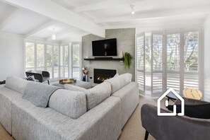 A modern living room with a large sectional sofa, a flat-screen TV, a fireplace, and large windows with blinds.