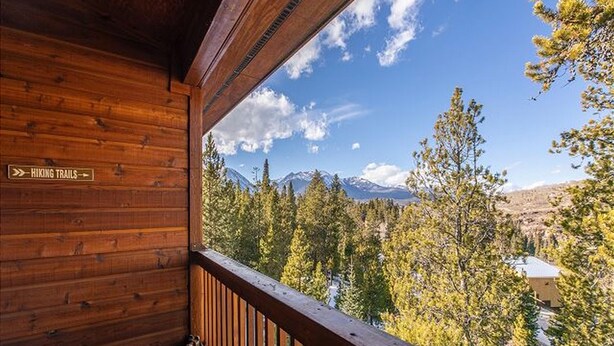 A wooden cabin with a balcony overlooking a forested mountain area and a sign indicating hiking trails. - Lookout Ridge Townhomes #113 by Summit County Mountain Retreats (Dillon)