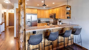 A modern kitchen with wooden cabinets, a stainless steel refrigerator, and a central island with bar stools. - Lookout Ridge Townhomes #113 by Summit County Mountain Retreats (Dillon)