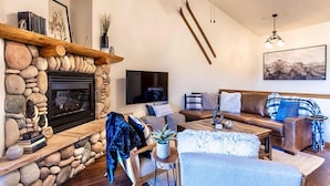 A living room with a stone fireplace, a ceiling fan, a flat-screen TV, a sofa with cushions, and a coffee table. - Lookout Ridge Townhomes #113 by Summit County Mountain Retreats (Dillon)