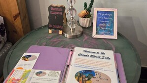 A small table with a lamp, a sign indicating maximum occupancy, a framed quote, and a welcome booklet for The Blank Canvas Artist Suites. - The Monet Suite is one of our four rooms. (Hermann)
