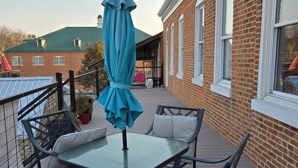 An outdoor patio with a glass table and metal chairs, a blue umbrella, and a brick building in the background. - The Monet Suite is one of our four rooms. (Hermann)