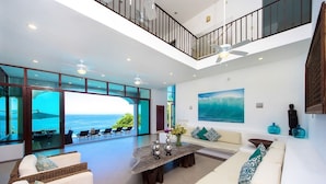 A spacious living area with a large window overlooking the ocean, a high ceiling with a fan, and a modern design with a wooden coffee table and white sofas. - PAY 6 STAY 7 NIGHTS until Oct 31. Luxury Villa at Conchas Chinas w/ Ocean Views (Puerto Vallarta)