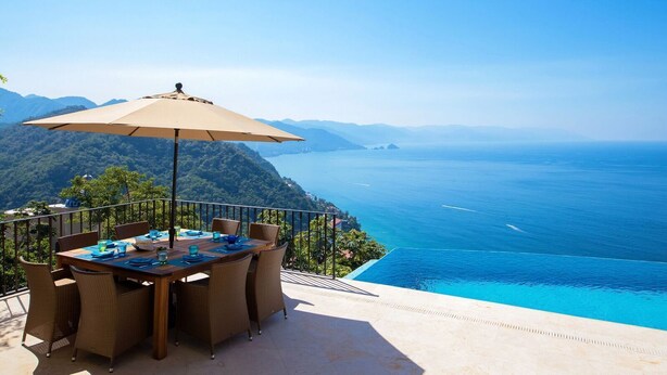 A dining table with chairs and a table umbrella on a balcony overlooking a clear blue sea and a mountainous landscape. - PAY 6 STAY 7 NIGHTS until Oct 31. Luxury Villa at Conchas Chinas w/ Ocean Views (Puerto Vallarta)